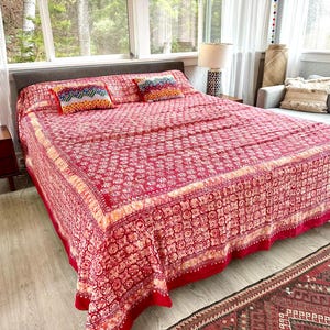 May include: A red and white patterned bedspread covers a bed, with two decorative pillows. The bedspread has a detailed floral design and a contrasting border. The room has a neutral color palette with natural light.