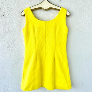 Pu&ograve; includere: Un abito giallo brillante senza maniche con scollo rotondo e tessuto strutturato. L'abito &egrave; appeso a un appendiabiti di legno.
