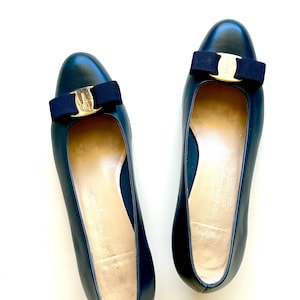 May include: A pair of dark navy blue leather pumps featuring a rounded toe and a dark navy blue grosgrain ribbon bow on the vamp. Each shoe is accented with a gold-tone metal hardware piece centered on the bow, featuring engraved branding.