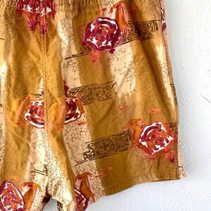 May include: Tan and brown shorts with an all-over floral and geometric pattern. The shorts have an elastic waistband and a relaxed fit. The floral design features red and white accents, adding a pop of color to the garment.