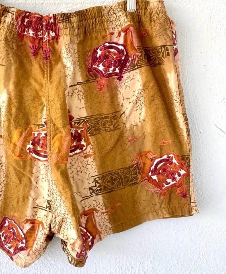 May include: Tan and brown shorts with an all-over floral and geometric pattern. The shorts have an elastic waistband and a relaxed fit. The floral design features red and white accents, adding a pop of color to the garment.