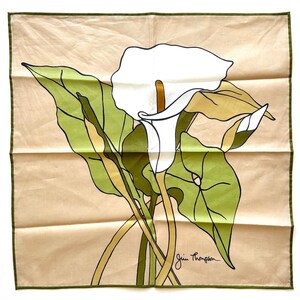 May include: A square scarf featuring a white calla lily with green leaves and stems against a tan background. The scarf has a green border and the signature "Jim Thompson" in the bottom right corner. The floral design is a stylized illustration.