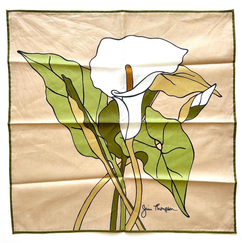 May include: A square scarf featuring a white calla lily with green leaves and stems against a tan background. The scarf has a green border and the signature "Jim Thompson" in the bottom right corner. The floral design is a stylized illustration.