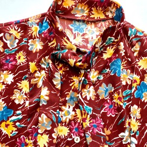 Pode incluir: Uma blusa marrom com estampa floral com flores amarelas, azuis e rosas. A blusa tem um la&ccedil;o no pesco&ccedil;o e um fecho de bot&atilde;o.