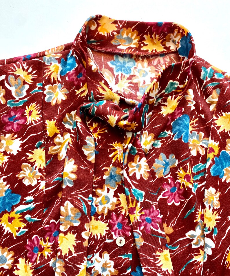 Pode incluir: Uma blusa marrom com estampa floral com flores amarelas, azuis e rosas. A blusa tem um la&ccedil;o no pesco&ccedil;o e um fecho de bot&atilde;o.
