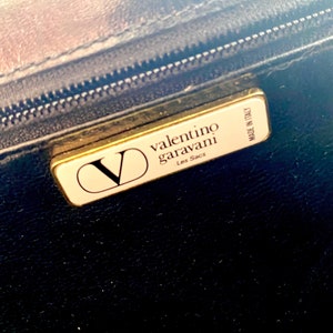 Op de afbeelding: Een goudkleurig label met de tekst "valentino garavani Les Sacs" en "MADE IN ITALY" op een zwarte achtergrond.