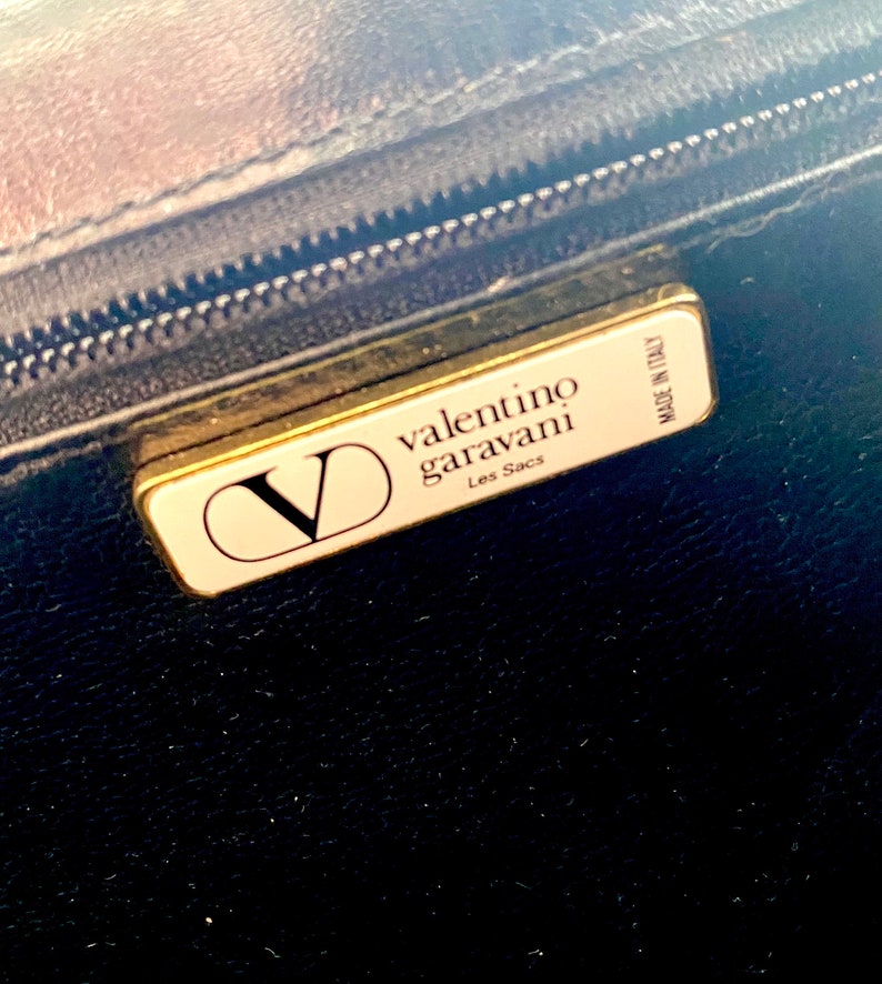 Op de afbeelding: Een goudkleurig label met de tekst "valentino garavani Les Sacs" en "MADE IN ITALY" op een zwarte achtergrond.