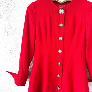 Puede incluir: Una blazer roja brillante, de manga larga, con cuello mandar&iacute;n y botones plateados. La blazer est&aacute; colgada de una percha vintage. Las mangas tienen un detalle de pu&ntilde;o doblado. Los botones son redondos y tienen un dise&ntilde;o decorativo.