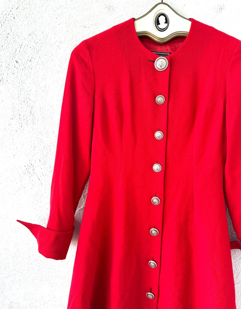 Puede incluir: Una blazer roja brillante, de manga larga, con cuello mandar&iacute;n y botones plateados. La blazer est&aacute; colgada de una percha vintage. Las mangas tienen un detalle de pu&ntilde;o doblado. Los botones son redondos y tienen un dise&ntilde;o decorativo.