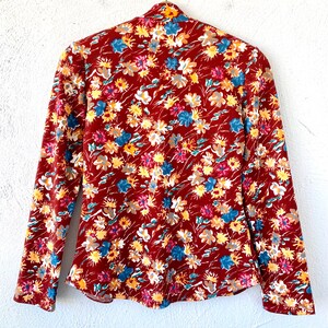 Pode incluir: Uma blusa de manga comprida cor ferrugem com estampa floral de pequenas flores azuis, amarelas e brancas. A blusa tem um decote alto e uma silhueta ajustada.