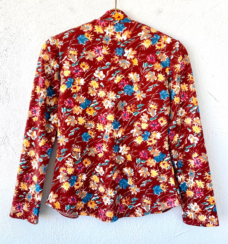 Pode incluir: Uma blusa de manga comprida cor ferrugem com estampa floral de pequenas flores azuis, amarelas e brancas. A blusa tem um decote alto e uma silhueta ajustada.