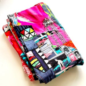 May include: A stack of colorful patchwork fabric items. The top layer features a vibrant pink floral pattern, while the layers below display a mix of geometric shapes, comic book-style illustrations, and skull motifs. The edges are neatly sewn with a dark blue thread.