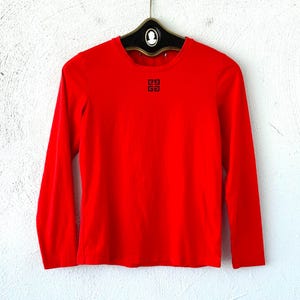 Pode incluir: Uma camisa vermelha de manga comprida com um logotipo preto na frente. O logotipo é um quadrado com quatro quadrados menores dentro.