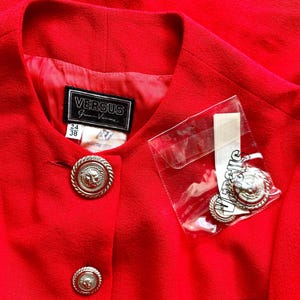 Puede incluir: Una chaqueta roja brillante con botones plateados con cabeza de le&oacute;n. La chaqueta presenta una etiqueta Versus Gianni Versace y botones adicionales en una bolsa de pl&aacute;stico transparente.