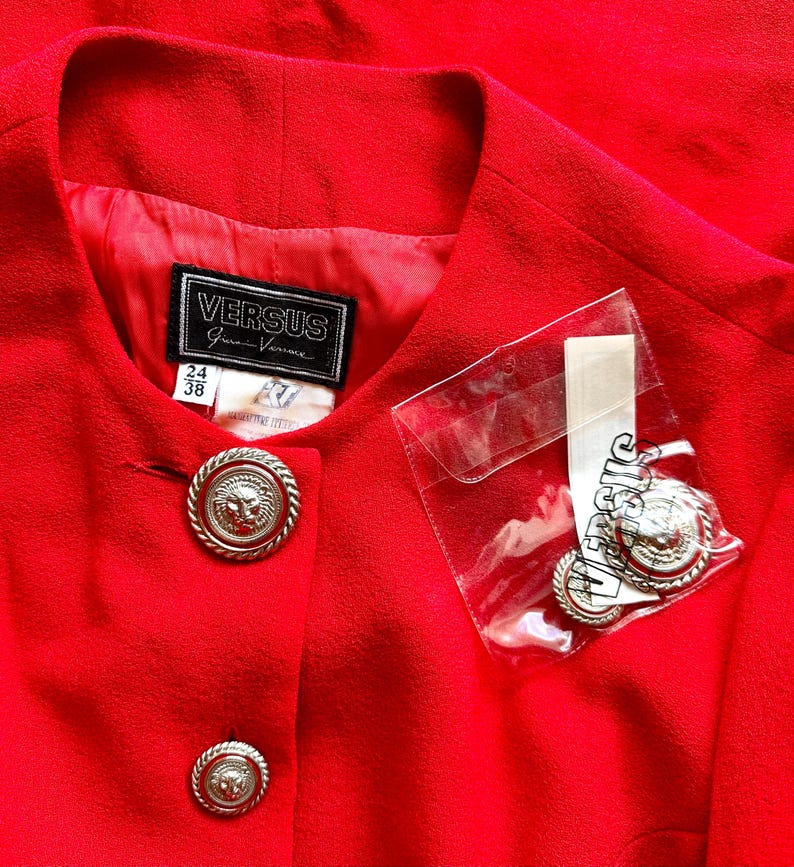 Puede incluir: Una chaqueta roja brillante con botones plateados con cabeza de le&oacute;n. La chaqueta presenta una etiqueta Versus Gianni Versace y botones adicionales en una bolsa de pl&aacute;stico transparente.