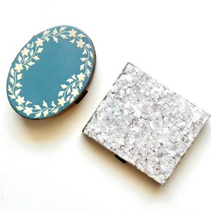 May include: Two decorative boxes. One is oval, teal with a white floral border. The other is square, covered in a shimmering, glittery material. Both boxes are closed and set against a white background.