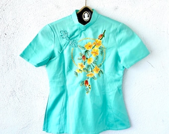 May include: A light blue short-sleeved blouse with a mandarin collar and a floral embroidery design. The embroidery features yellow flowers and green leaves in a circular pattern.