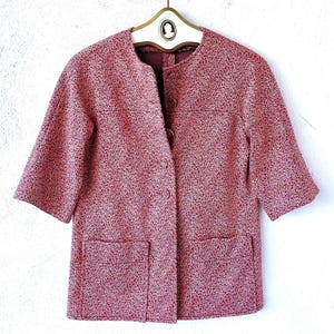 May include: A short-sleeved jacket in a speckled red and white textured fabric. The jacket features a round neckline, a front button closure with four visible buttons, and two large square patch pockets at the bottom front hem.