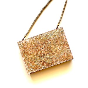 May include: A rectangular clutch with a gold-colored chain strap. The body of the bag is covered in a textured, gold and tan pattern, giving it a shimmering appearance. The bag is set against a white background.
