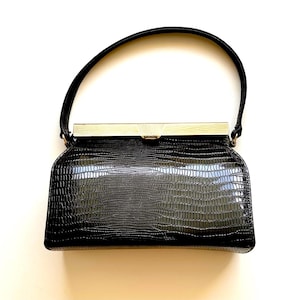 May include: A black handbag with a glossy, textured lizard-skin pattern. The bag features a single arched handle and a rectangular gold-tone metal frame closure with a subtle sunburst engraving at the center of the clasp mechanism.
