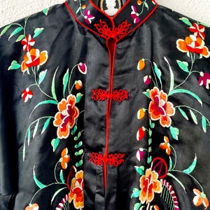 Puede incluir: Una bata de seda negra con ribetes rojos y bordados florales en tonos naranja, rojo, verde y blanco.