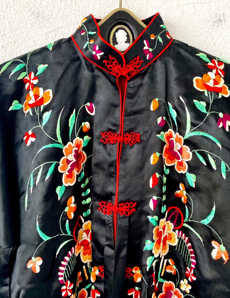 Puede incluir: Una bata de seda negra con ribetes rojos y bordados florales en tonos naranja, rojo, verde y blanco.