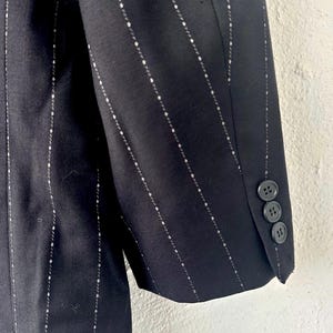 Peut inclure: Une manche de blazer noire orn&eacute;e d'un motif discret de fines rayures blanches compos&eacute;es de petits losanges r&eacute;p&eacute;titifs. Le poignet de la manche est agr&eacute;ment&eacute; de trois boutons ronds de couleur sombre align&eacute;s verticalement sur le bord ext&eacute;rieur.