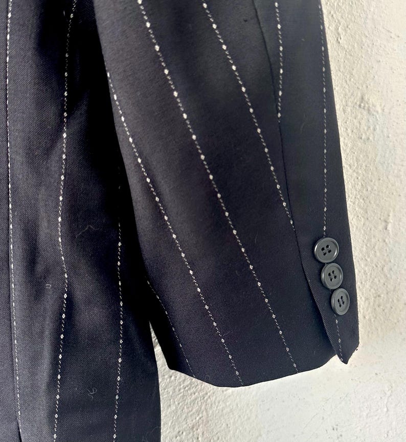 Peut inclure: Une manche de blazer noire orn&eacute;e d'un motif discret de fines rayures blanches compos&eacute;es de petits losanges r&eacute;p&eacute;titifs. Le poignet de la manche est agr&eacute;ment&eacute; de trois boutons ronds de couleur sombre align&eacute;s verticalement sur le bord ext&eacute;rieur.