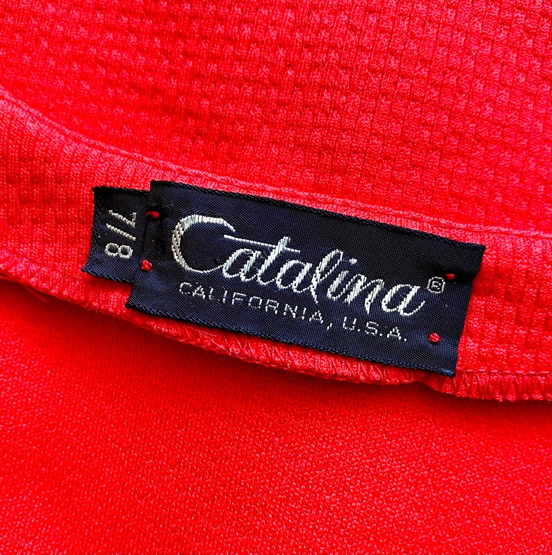 May include: Close-up of a bright red fabric with a textured surface. A navy blue label with the name "Catalina" in cursive script is attached. The label also includes "CALIFORNIA, U.S.A." and the size "7/8".