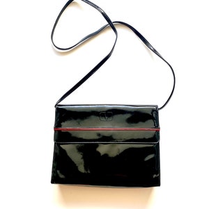 Op de afbeelding: Een zwarte crossbody tas van gelakt leer met een rode bies en een klein logo aan de voorkant.