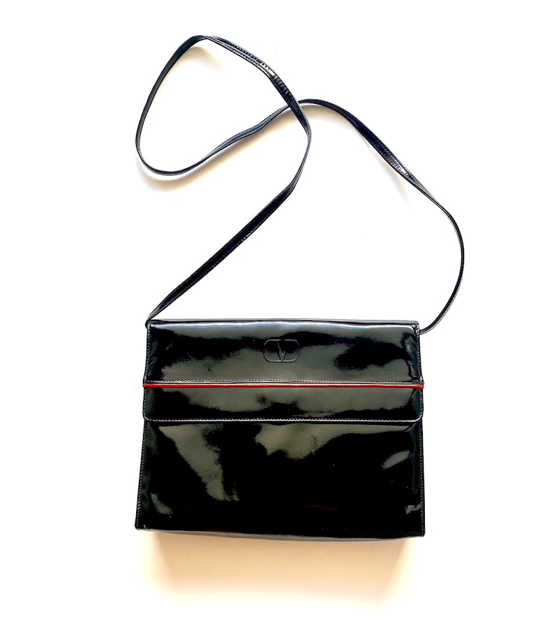 Op de afbeelding: Een zwarte crossbody tas van gelakt leer met een rode bies en een klein logo aan de voorkant.