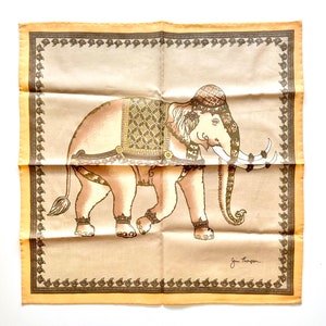 May include: A tan and brown decorative square scarf featuring an illustrated elephant with ornate detailing. The elephant is adorned with a patterned saddle and head covering. The scarf has a decorative border and is signed "Jen Thompson".