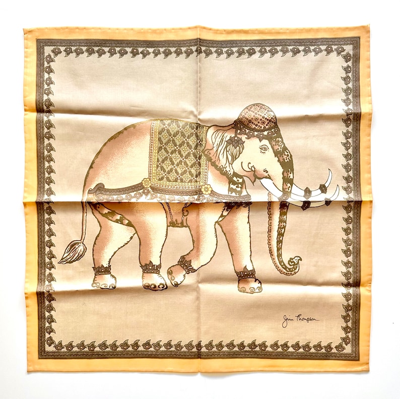 May include: A tan and brown decorative square scarf featuring an illustrated elephant with ornate detailing. The elephant is adorned with a patterned saddle and head covering. The scarf has a decorative border and is signed "Jen Thompson".