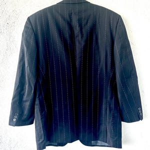 Peut inclure: Un blazer bleu marine fonc&eacute; orn&eacute; d'un motif &agrave; fines rayures verticales blanches. Le v&ecirc;tement pr&eacute;sente des manches longues avec trois petits boutons sur chaque poignet, un col &agrave; revers crant&eacute; et des &eacute;paules structur&eacute;es, vu de dos.