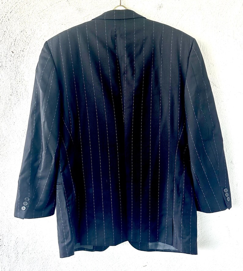 Peut inclure: Un blazer bleu marine fonc&eacute; orn&eacute; d'un motif &agrave; fines rayures verticales blanches. Le v&ecirc;tement pr&eacute;sente des manches longues avec trois petits boutons sur chaque poignet, un col &agrave; revers crant&eacute; et des &eacute;paules structur&eacute;es, vu de dos.