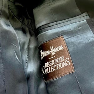 Peut inclure: L'int&eacute;rieur d'un blazer gris anthracite fonc&eacute; pr&eacute;sentant une &eacute;tiquette en tissu rectangulaire marron cousue sur la doublure. L'&eacute;tiquette affiche le texte brod&eacute; en blanc Neiman Marcus DESIGNER COLLECTIONS avec une ligne d&eacute;corative au-dessus du mot DESIGNER.