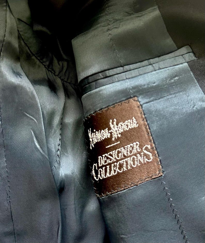 Peut inclure: L'int&eacute;rieur d'un blazer gris anthracite fonc&eacute; pr&eacute;sentant une &eacute;tiquette en tissu rectangulaire marron cousue sur la doublure. L'&eacute;tiquette affiche le texte brod&eacute; en blanc Neiman Marcus DESIGNER COLLECTIONS avec une ligne d&eacute;corative au-dessus du mot DESIGNER.