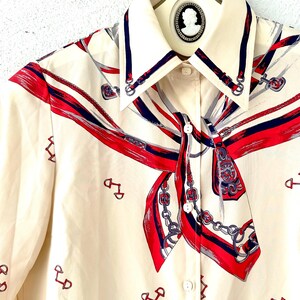 May include: A cream-colored button-up shirt with a long-sleeve design. The shirt features a red, blue, and white patterned design of a scarf with chains and horse bits.