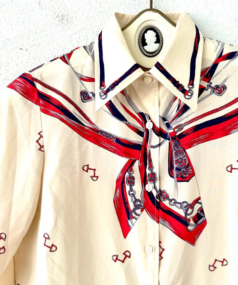 May include: A cream-colored button-up shirt with a long-sleeve design. The shirt features a red, blue, and white patterned design of a scarf with chains and horse bits.