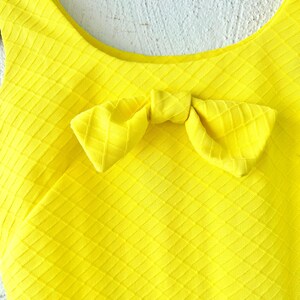 Pu&ograve; includere: Un top giallo brillante senza maniche con un dettaglio a fiocco sul collo. Il top ha un motivo testurizzato a forma di diamante.