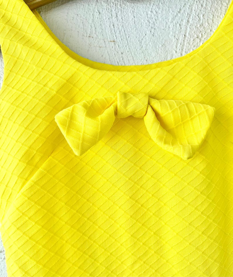 Pu&ograve; includere: Un top giallo brillante senza maniche con un dettaglio a fiocco sul collo. Il top ha un motivo testurizzato a forma di diamante.