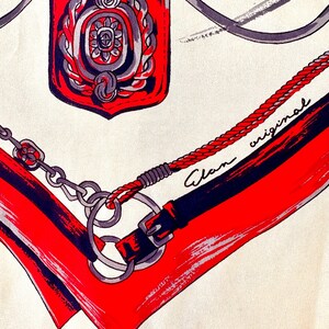 May include: A red, white, and black patterned scarf with a silver chain and a red and silver emblem. The scarf has the text "Alan original" written on it.