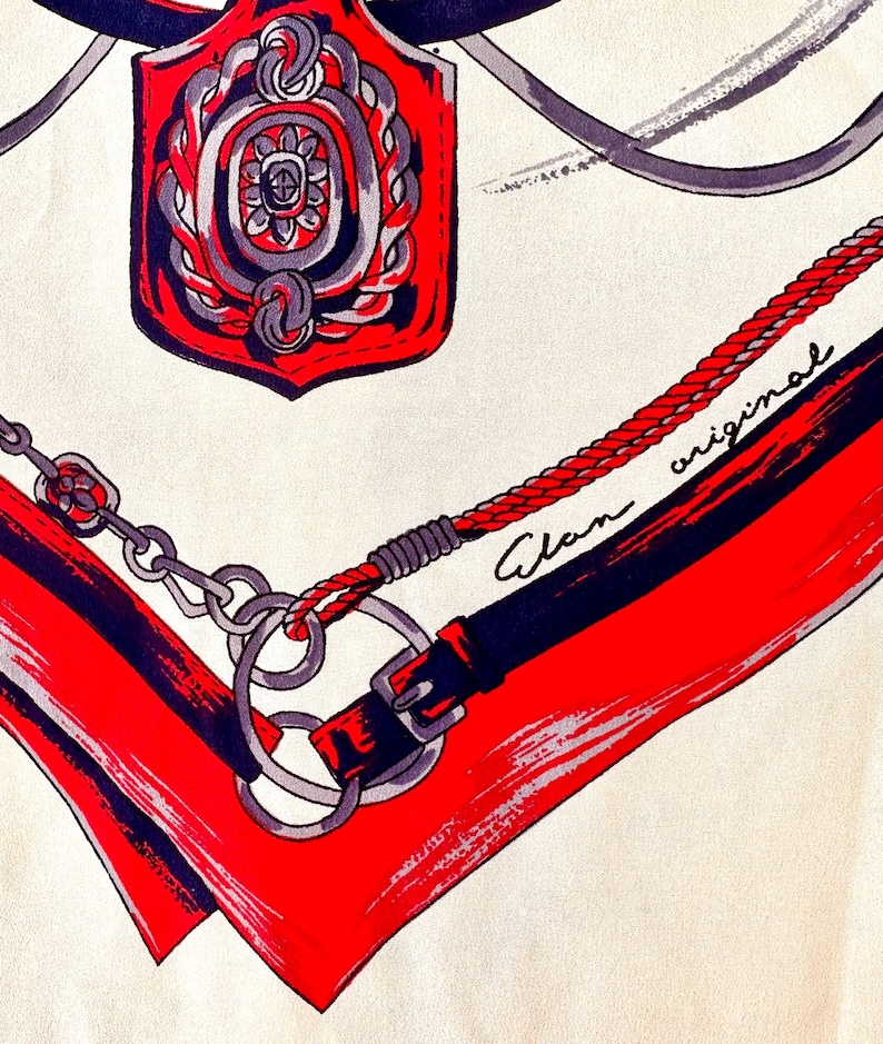 May include: A red, white, and black patterned scarf with a silver chain and a red and silver emblem. The scarf has the text "Alan original" written on it.