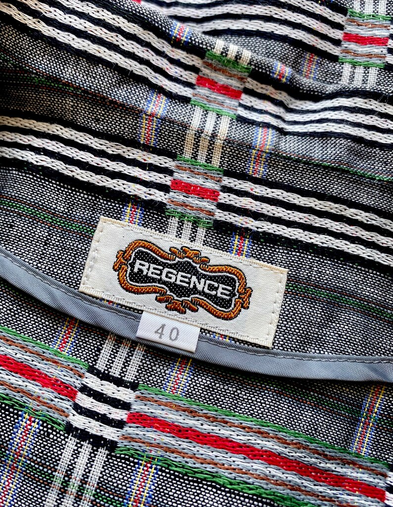 May include: A garment featuring a black, white, red, and green woven plaid pattern. A rectangular white fabric label is stitched to the interior, displaying the text REGENCE in black letters within a decorative gold border, with a size 40 tag below.