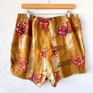 May include: Tan shorts with an elastic waistband, featuring a repeating pattern of burgundy roses and geometric shapes. The shorts are hanging on a wooden hanger against a white background.