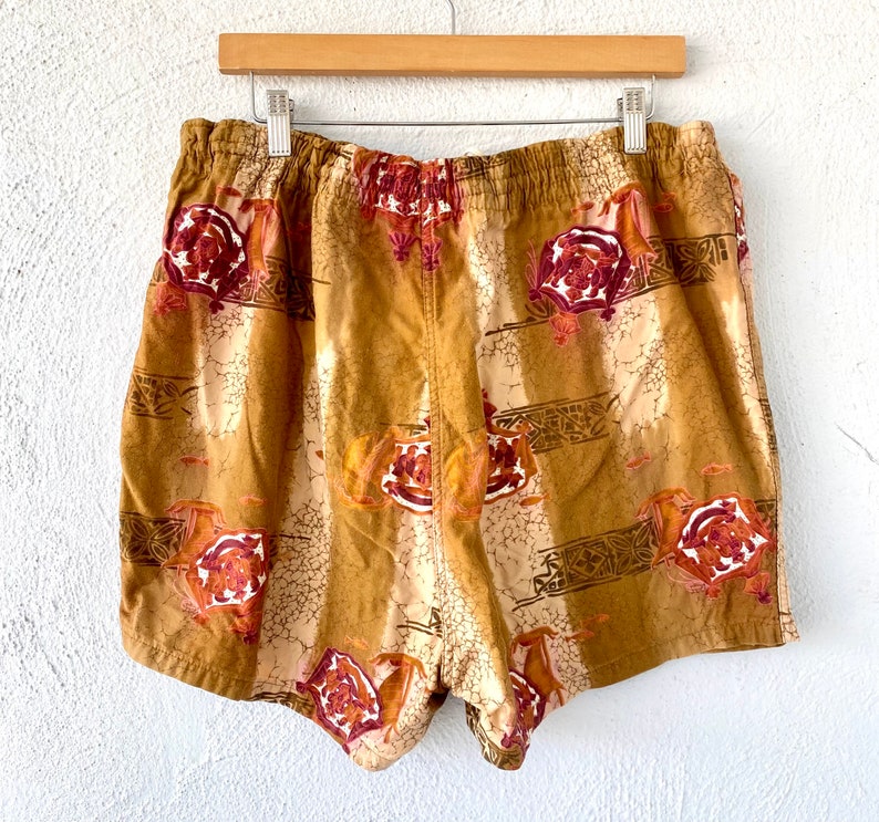 May include: Tan shorts with an elastic waistband, featuring a repeating pattern of burgundy roses and geometric shapes. The shorts are hanging on a wooden hanger against a white background.