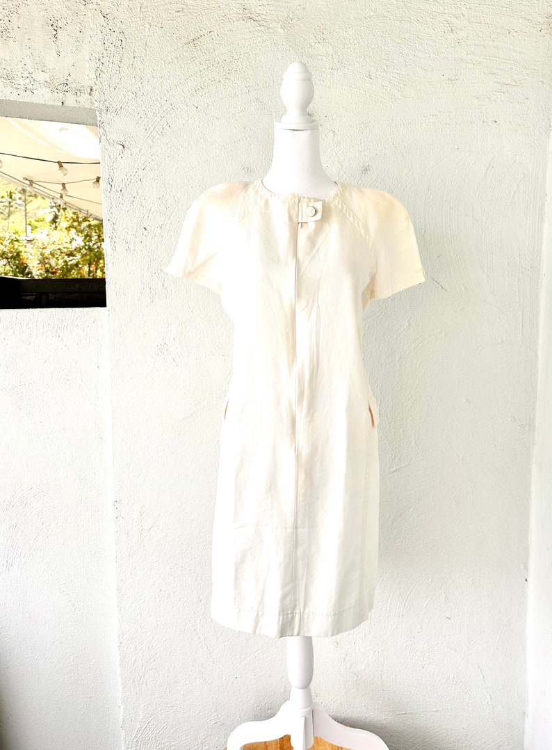 May include: A white linen dress with short sleeves and a button closure at the neck. The dress has two pockets on the front and a fitted silhouette.