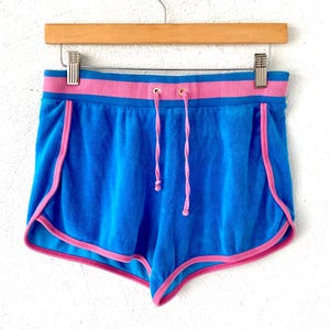 Puede incluir: Shorts de rizo azul con ribete rosa y cintura rosa. Los shorts tienen cierre de cordón y cuelgan de una percha de madera. Los shorts son de un color azul vibrante.