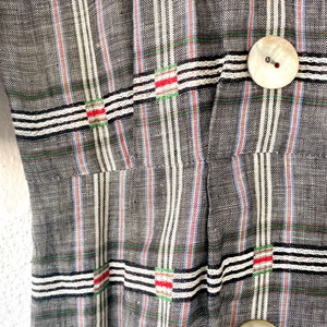 May include: A grey fabric garment featuring a vertical and horizontal striped pattern in white, black, red, and green. The piece includes two large, round, off-white buttons with a pearlescent finish, positioned vertically along the right side.