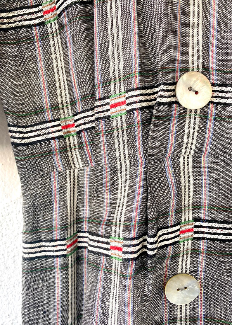 May include: A grey fabric garment featuring a vertical and horizontal striped pattern in white, black, red, and green. The piece includes two large, round, off-white buttons with a pearlescent finish, positioned vertically along the right side.